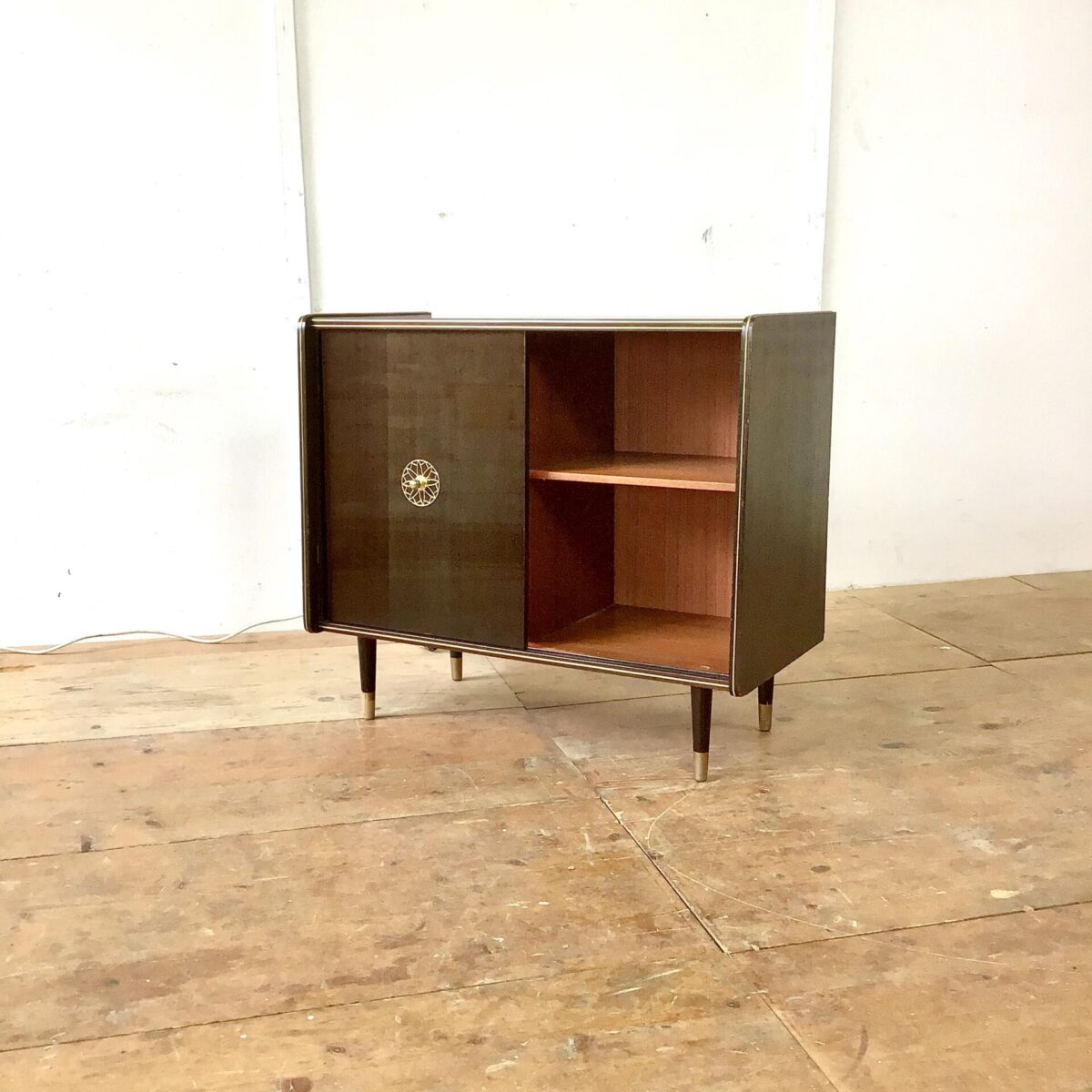 Vintage Sideboard 84.5cm mal 41cm Höhe 74cm. Hinter der Glas Schiebetür ist das Möbel mit goldigem Stoff ausgekleidet. Am Boden hat’s ein Spiegel und von oben bringt eine kleine Lampe Licht in die Vitrine. Die rechte Hälfte der Kommode bietet etwas stau Raum mit einem Tablar. Der Messing Türgriff mit der Sonnen artigen Verzierung wiederholt sich bei der Glastüre, dort ist es ins hinterglas eingefräst. Oben auf dem Sideboard hat’s ein paar Kratzer im Lack. Bei Füssen ist die untere, goldige Farbe teilweise abgeblättert. Ansonsten in stabilem funktionalen Zustand. 