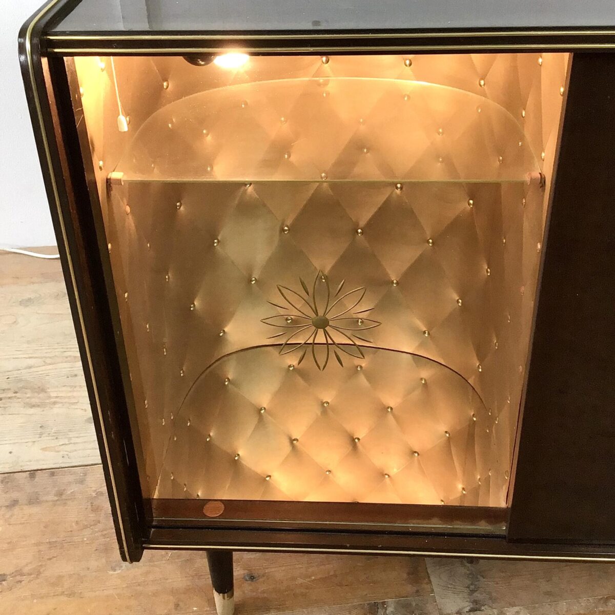 Vintage Sideboard 84.5cm mal 41cm Höhe 74cm. Hinter der Glas Schiebetür ist das Möbel mit goldigem Stoff ausgekleidet. Am Boden hat’s ein Spiegel und von oben bringt eine kleine Lampe Licht in die Vitrine. Die rechte Hälfte der Kommode bietet etwas stau Raum mit einem Tablar. Der Messing Türgriff mit der Sonnen artigen Verzierung wiederholt sich bei der Glastüre, dort ist es ins hinterglas eingefräst. Oben auf dem Sideboard hat’s ein paar Kratzer im Lack. Bei Füssen ist die untere, goldige Farbe teilweise abgeblättert. Ansonsten in stabilem funktionalen Zustand. 