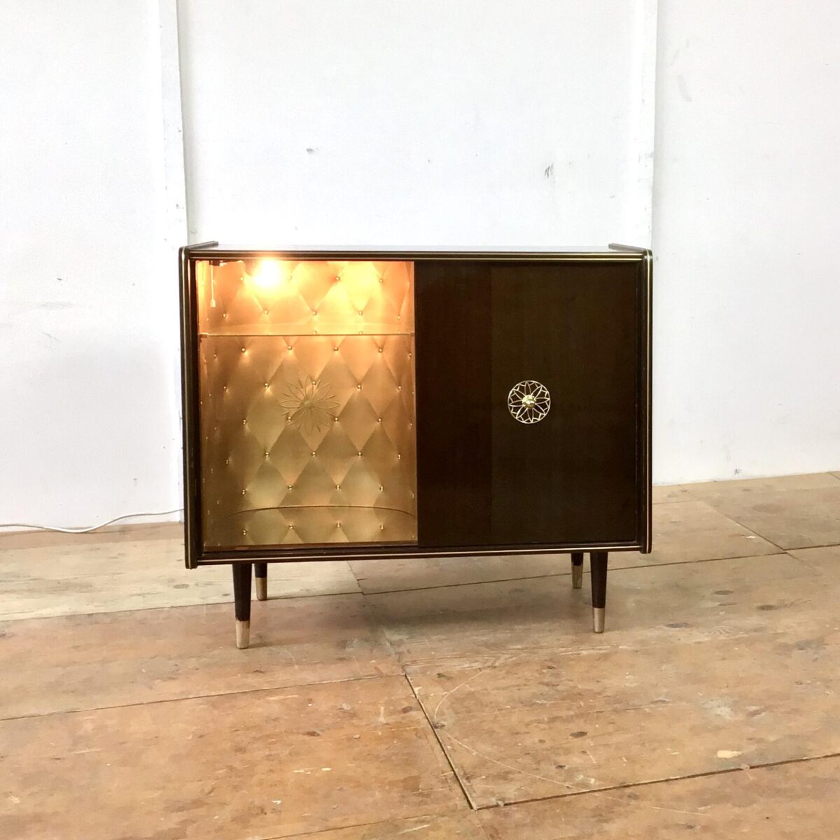 Vintage Sideboard 84.5cm mal 41cm Höhe 74cm. Hinter der Glas Schiebetür ist das Möbel mit goldigem Stoff ausgekleidet. Am Boden hat’s ein Spiegel und von oben bringt eine kleine Lampe Licht in die Vitrine. Die rechte Hälfte der Kommode bietet etwas stau Raum mit einem Tablar. Der Messing Türgriff mit der Sonnen artigen Verzierung wiederholt sich bei der Glastüre, dort ist es ins hinterglas eingefräst. Oben auf dem Sideboard hat’s ein paar Kratzer im Lack. Bei Füssen ist die untere, goldige Farbe teilweise abgeblättert. Ansonsten in stabilem funktionalen Zustand. 