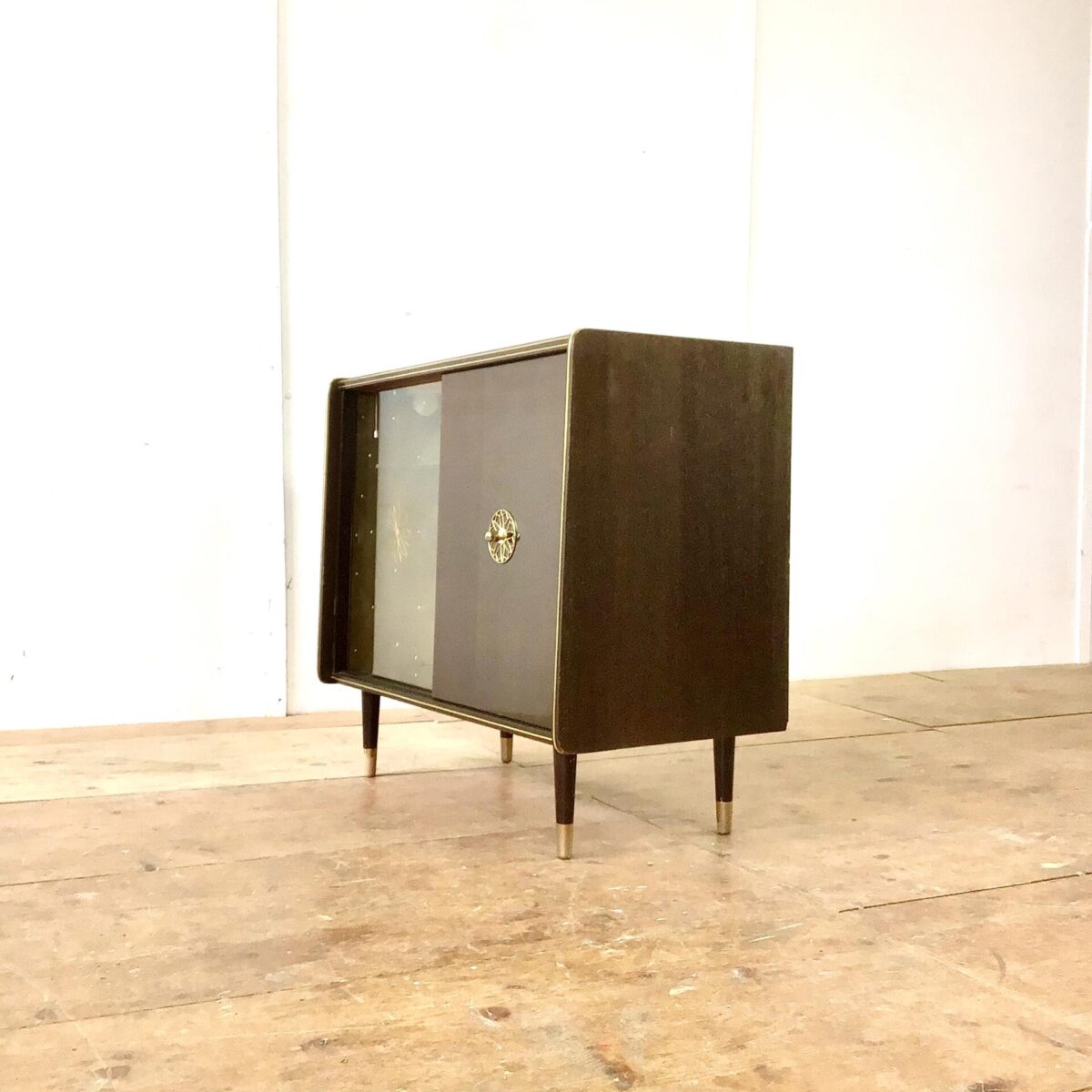 Vintage Sideboard 84.5cm mal 41cm Höhe 74cm. Hinter der Glas Schiebetür ist das Möbel mit goldigem Stoff ausgekleidet. Am Boden hat’s ein Spiegel und von oben bringt eine kleine Lampe Licht in die Vitrine. Die rechte Hälfte der Kommode bietet etwas stau Raum mit einem Tablar. Der Messing Türgriff mit der Sonnen artigen Verzierung wiederholt sich bei der Glastüre, dort ist es ins hinterglas eingefräst. Oben auf dem Sideboard hat’s ein paar Kratzer im Lack. Bei Füssen ist die untere, goldige Farbe teilweise abgeblättert. Ansonsten in stabilem funktionalen Zustand. 